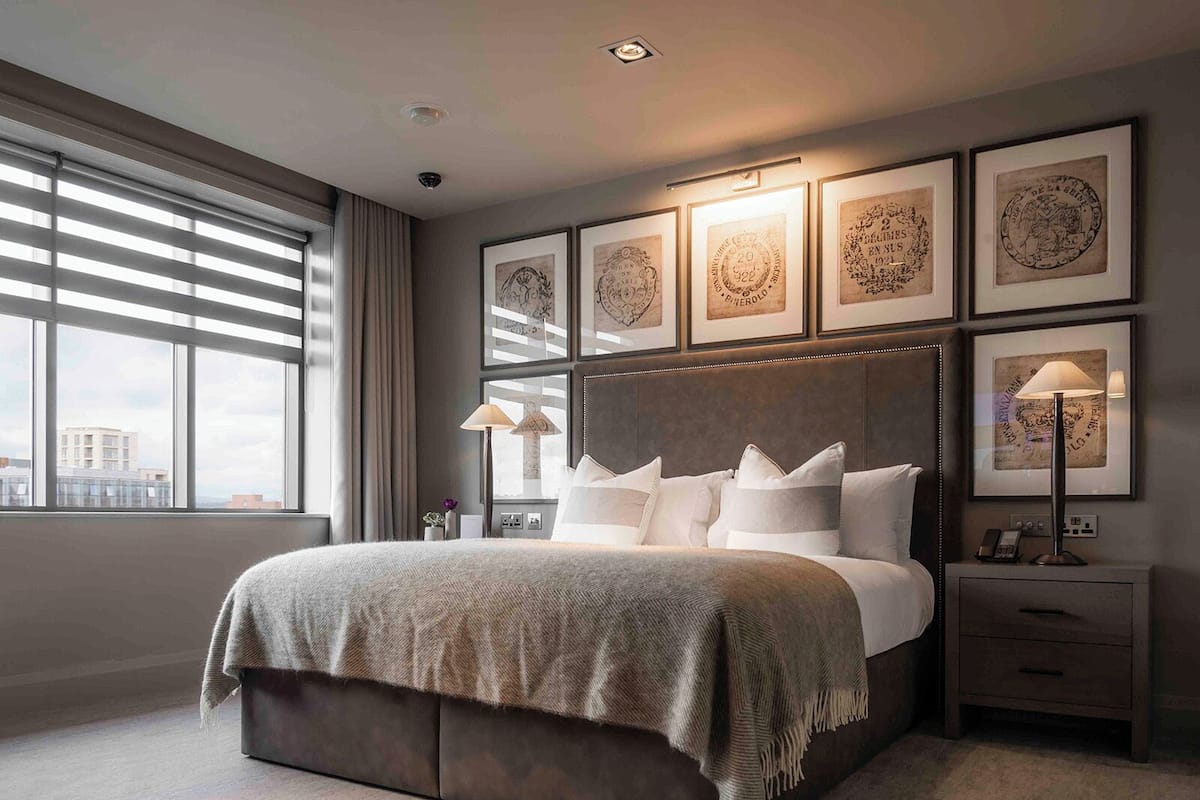 Et moderne hotelværelse med en stor seng, en sengegavl med indrammet kunst, et natbord med lampe og udsigt over byen gennem store vinduer.