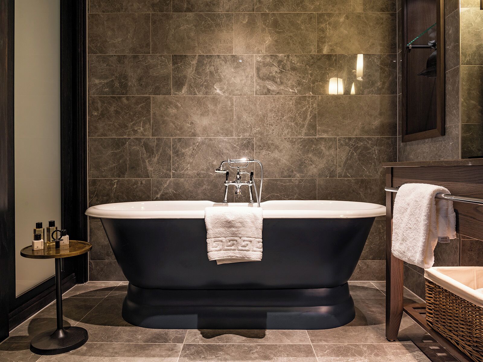Ein modernes Badezimmer mit einer freistehenden Badewanne, einem runden Tisch mit Pflegeprodukten und einem Korbstuhl.