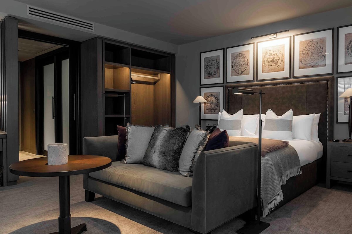 Et moderne hotelværelse med en grå sofa, et rundt sofabord, en seng med hvide sengetøj og en senelampe, samt indrammet kunst på væggen.