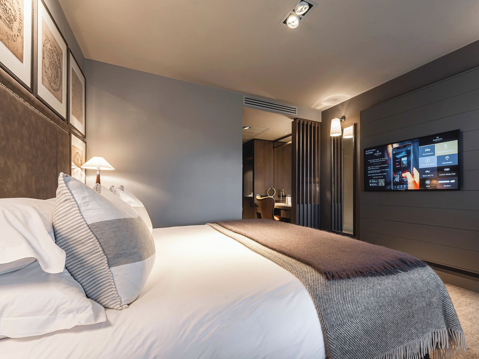 Quarto de hotel moderno com uma cama grande, uma escrivaninha e uma TV exibindo informações sobre o quarto.