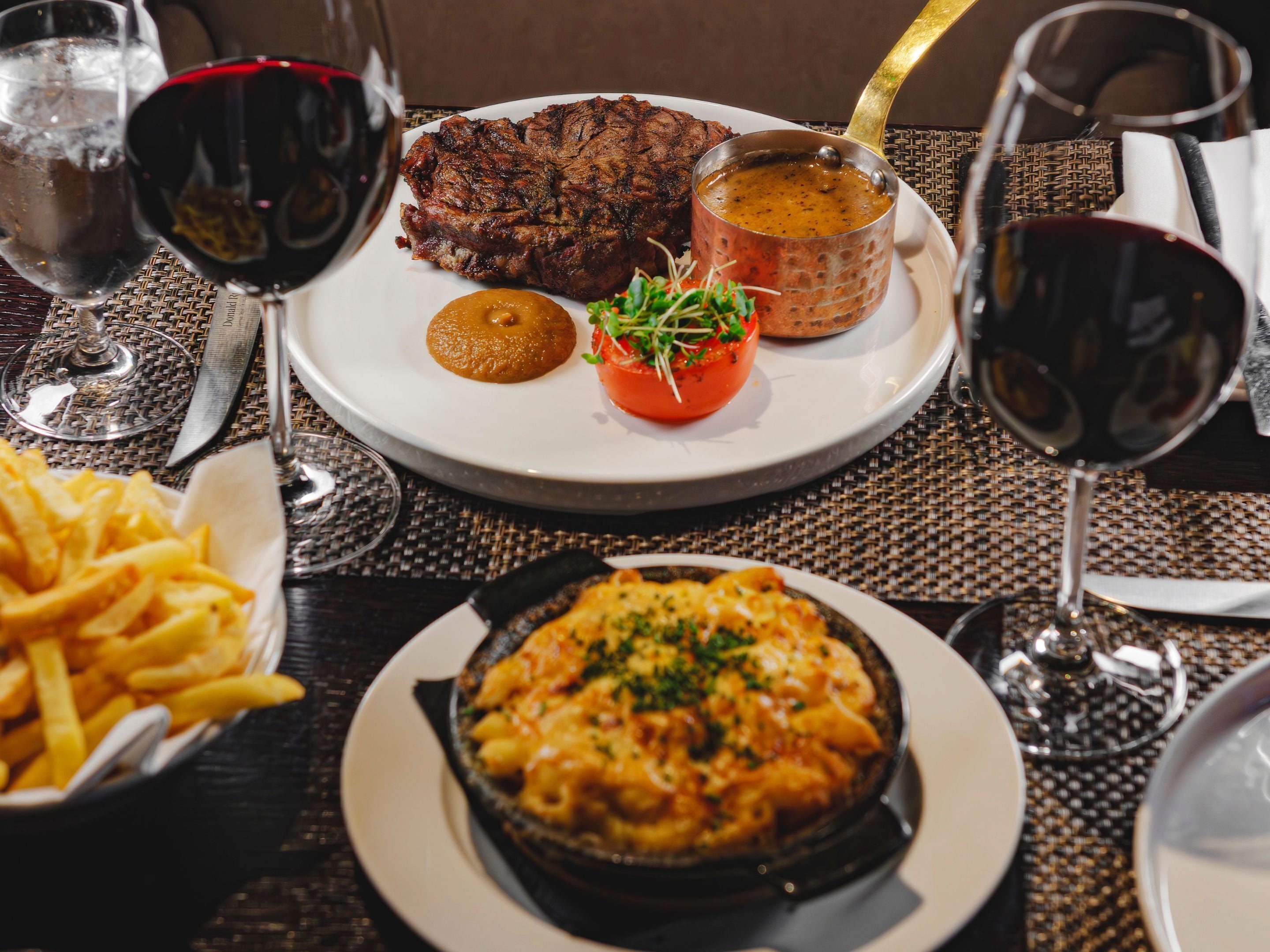Una mesa de comedor con un bistec, un bol de papas fritas, un plato de macarrones con queso y copas de vino tinto.