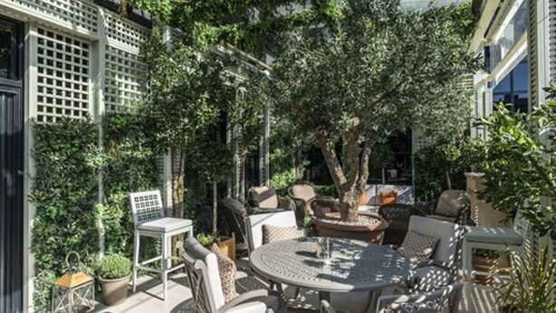 Eine Terrasse im Freien mit runden Tischen und Stühlen, umgeben von üppigem Grün und einem großen Baum.