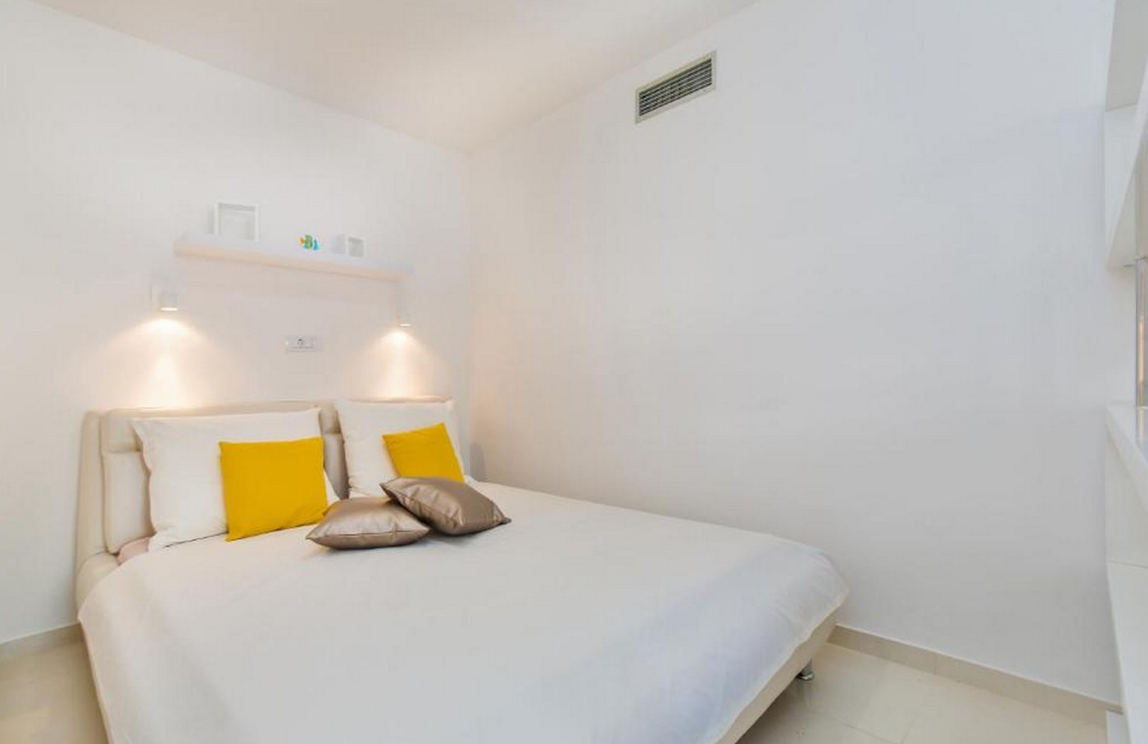 Une chambre à coucher avec un lit, des murs blancs, une étagère avec quelques objets et une grille d’aération sur le mur.