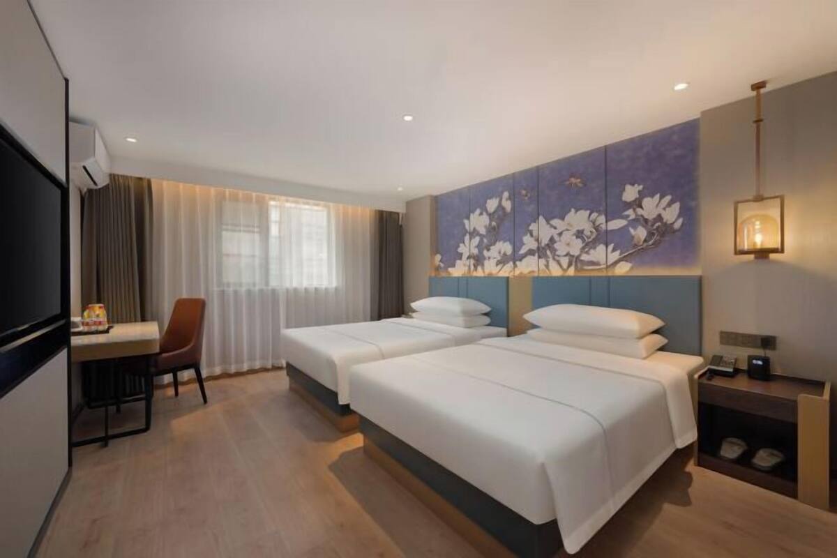 A hotel room with two beds, a desk, a chair, a TV, and a wall-mounted lamp.
