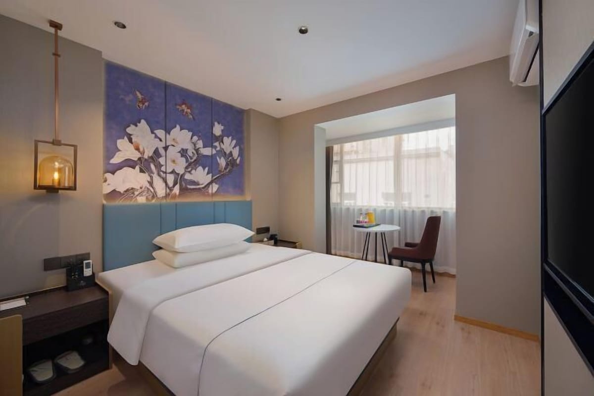 A hotel room with a large bed, a painting on the wall, a desk with a lamp, and a view of the outside through a window.