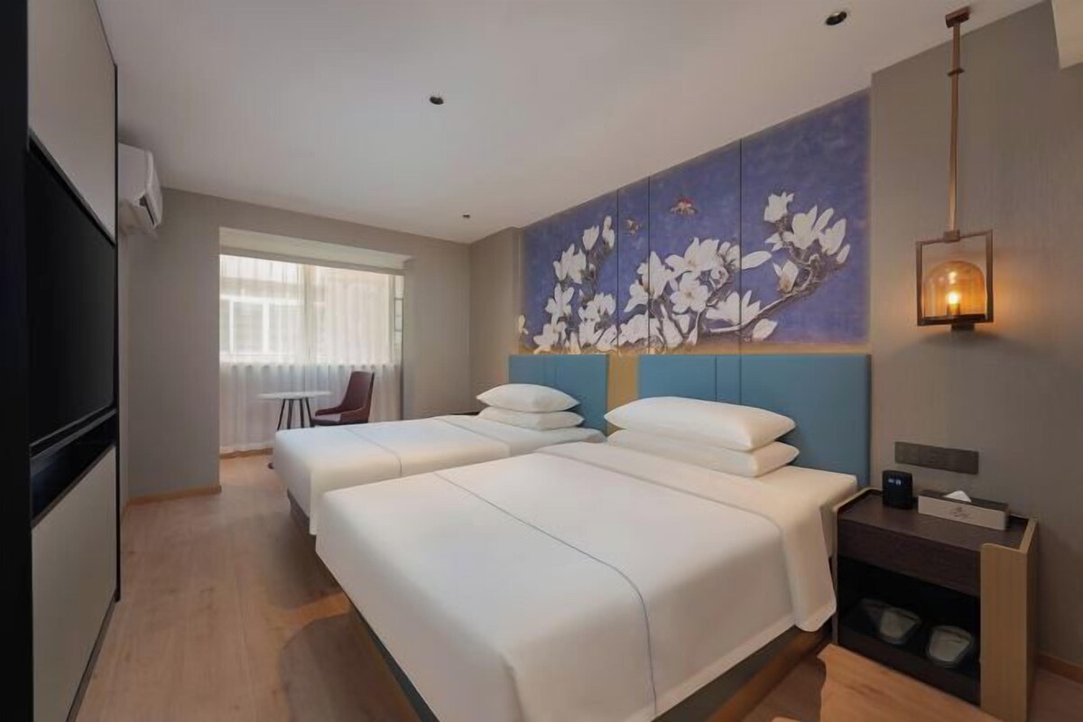 Quarto de hotel com duas camas, uma grande obra de arte floral na parede, uma mesinha de cabeceira com abajur e uma janela com cortinas.