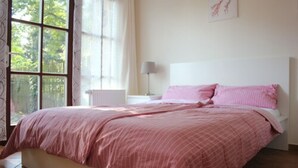 Une chambre avec une grande fenêtre, un lit avec une literie rose, une table de chevet avec une lampe et un tableau encadré au mur.