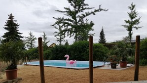 Une piscine extérieure avec un flotteur en forme de flamant rose, entourée de sable et de plantes en pot.