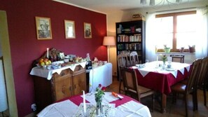 A dining area with a table set for two, a bookshelf, and a window with plants. - Pension & Restaurant Altjessen 57 (Pirna)