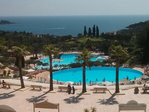 A large outdoor swimming pool surrounded by palm trees and benches, with a view of the sea in the background. - Residence Cap Esterel - Golf (Saint-Raphael)