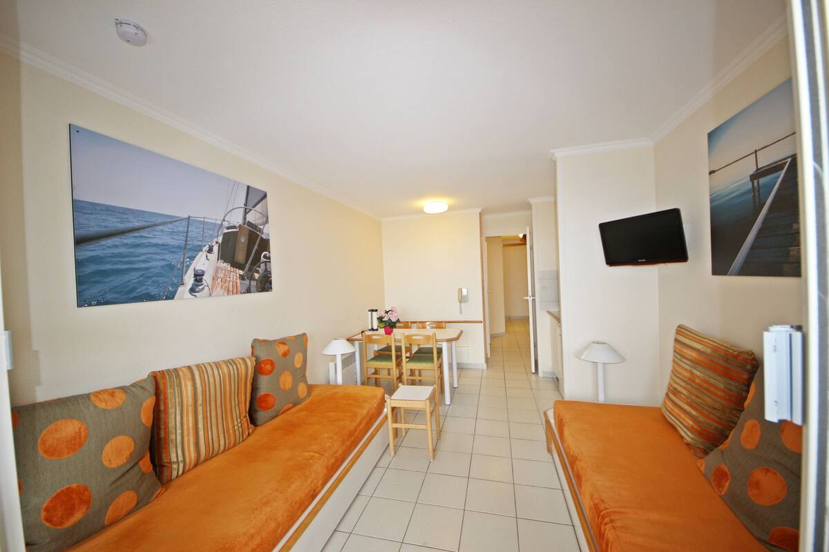 Il s’agit d’une pièce de taille modeste, aménagée avec deux canapés orange, une table à manger et des chaises. Deux grandes peintures ornent les murs, un téléviseur à écran plat est fixé au mur et un luminaire est suspendu au plafond.