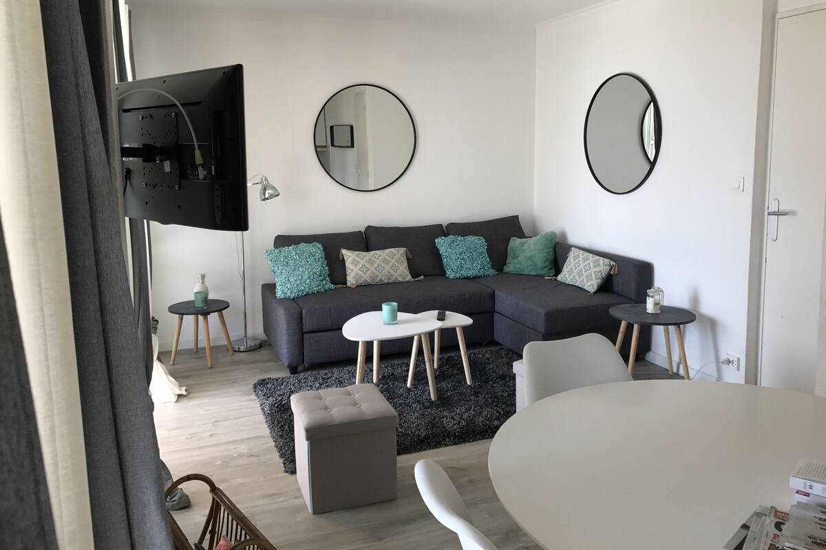 Un salon moderne avec un canapé d’angle gris, deux tables d’appoint, une table basse ronde et un téléviseur fixé au mur.