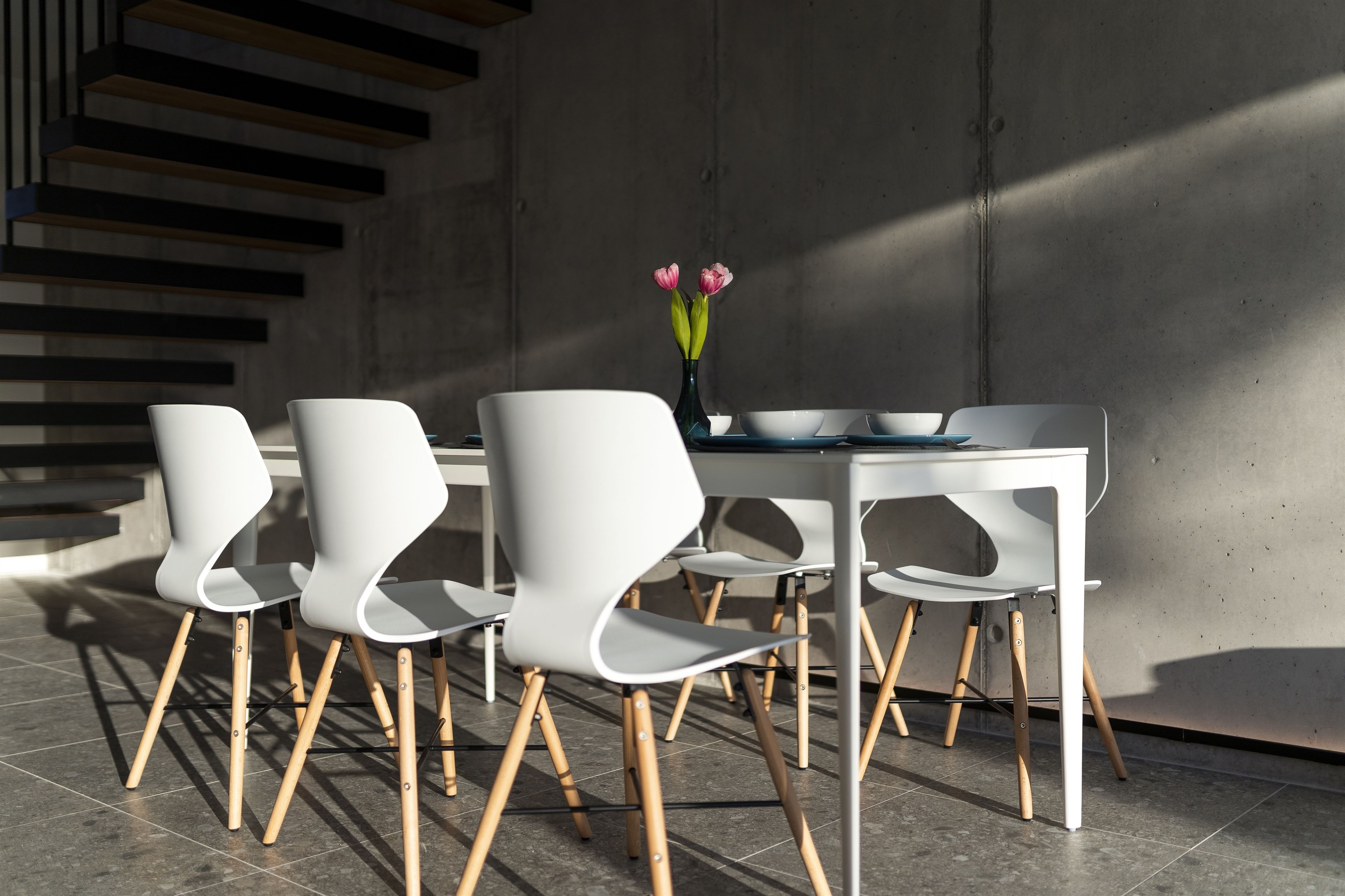 Uma sala de jantar moderna com uma mesa e cadeiras brancas, um vaso com flores e uma parede de concreto com uma escada de madeira.