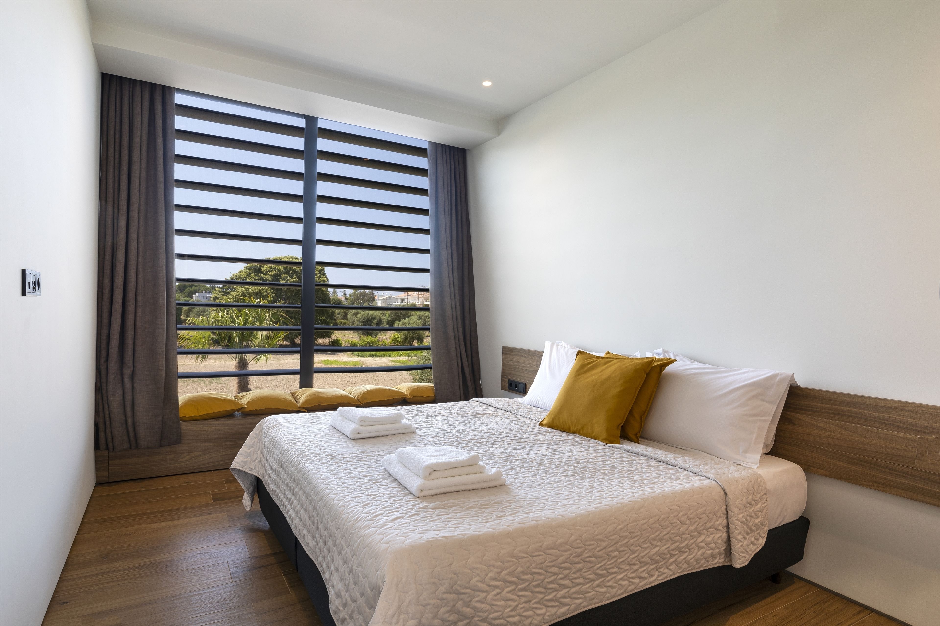 A modern hotel room with a large bed, a wooden headboard, and a view through floor-to-ceiling windows.