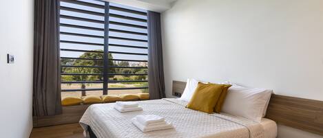 A modern hotel room with a large bed, a wooden headboard, and a view through floor-to-ceiling windows.