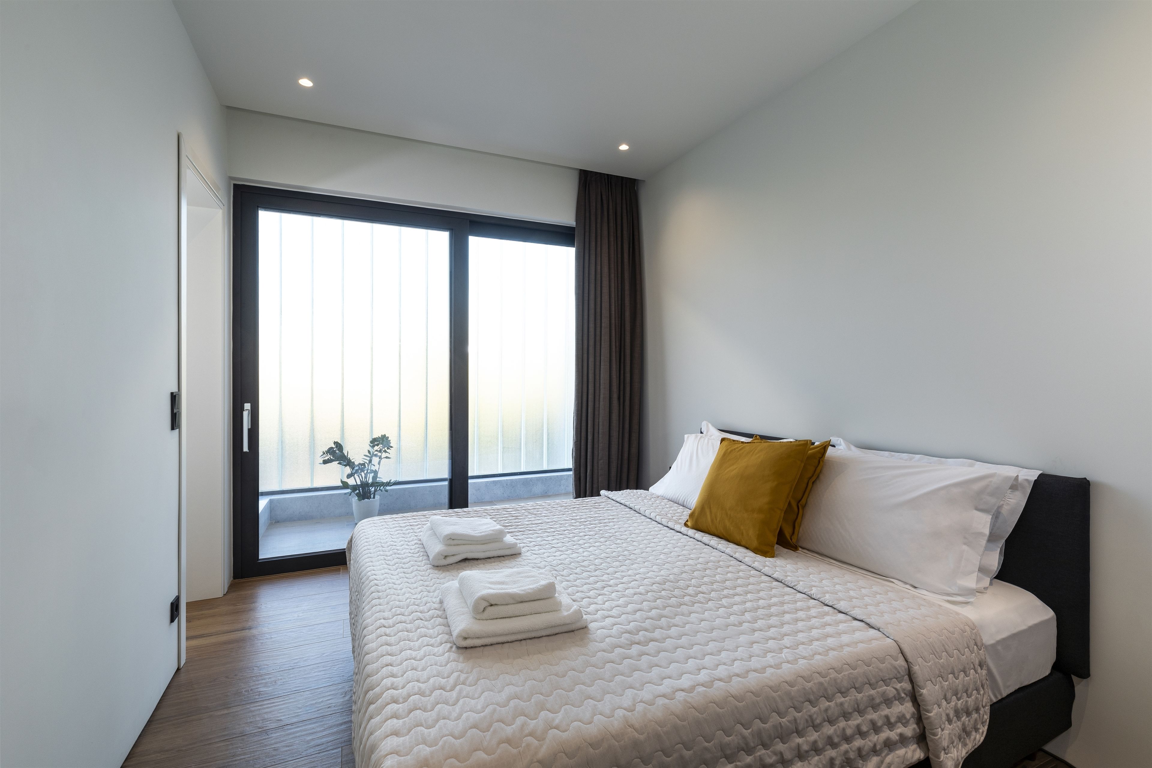 A modern hotel room with a large bed, a view of the city through floor-to-ceiling windows, and a balcony with a potted plant.