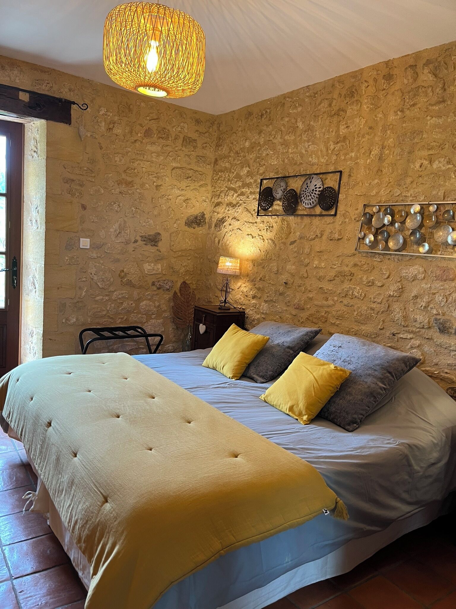 A bedroom with a stone wall, a bed with a yellow and grey duvet, a hanging lamp, and decorative wall art.
