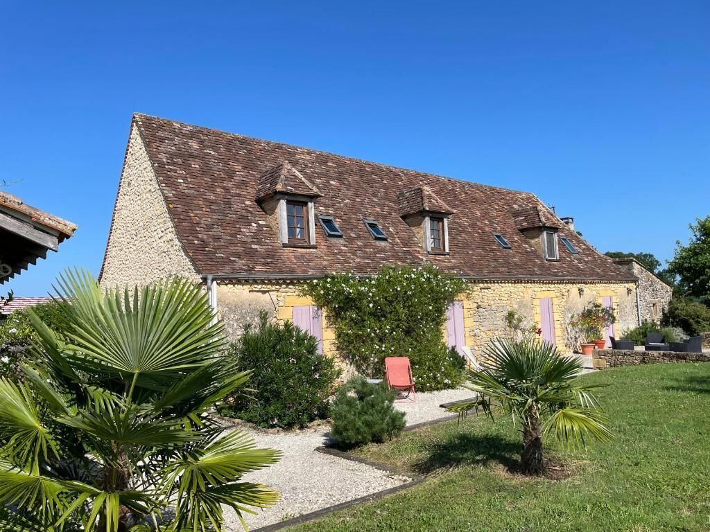 Une maison en pierre à deux étages, avec un toit en pente, un chemin de gravier et un petit jardin agrémenté de palmiers.