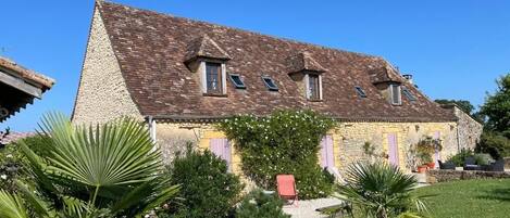 Une maison en pierre à deux étages, avec un toit en pente, un chemin de gravier et un petit jardin agrémenté de palmiers.