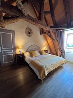 Quarto com teto de madeira, cama com colcha bege, duas luminárias de cabeceira e janela com cortinas.