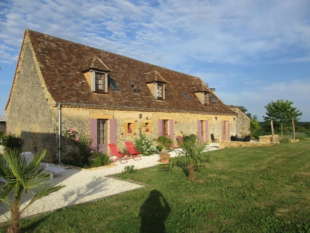 A stone house with a sloped roof, multiple windows, and a small garden area with red chairs and a palm tree.