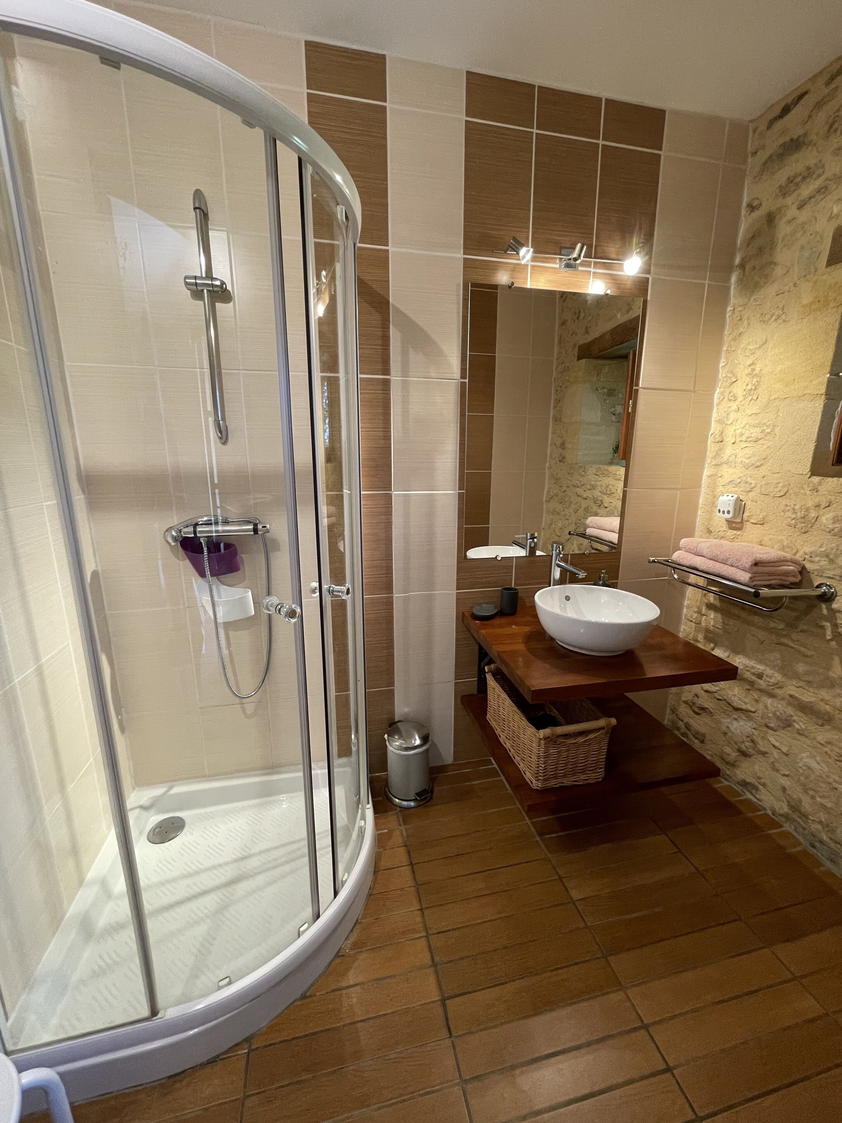 Un baño con ducha envuelta en vidrio, tocador de madera con lavamanos blanco y pared de piedra.