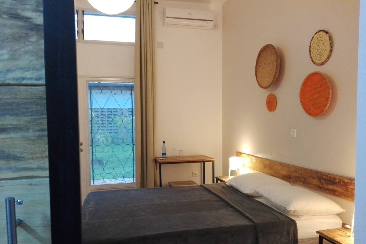 A hotel room with a bed, a bedside table with a lamp, a small table with a water bottle, and a window with a view of greenery.