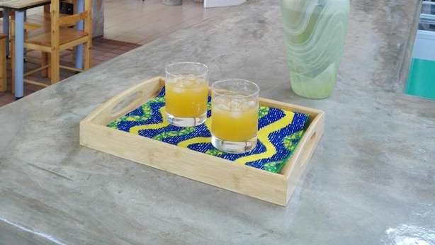 A dining area with a stone wall, a wooden table, chairs, a vase with yellow flowers, and two glasses of orange juice on a tray. - Urban Lodge (Bujumbura)