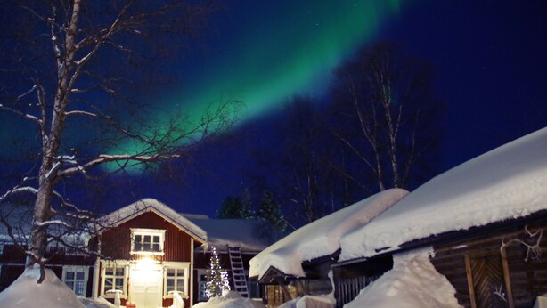 Una cabaña cubierta de nieve con la puerta iluminada, rodeada de montones de nieve, bajo un cielo nocturno con auroras boreales.