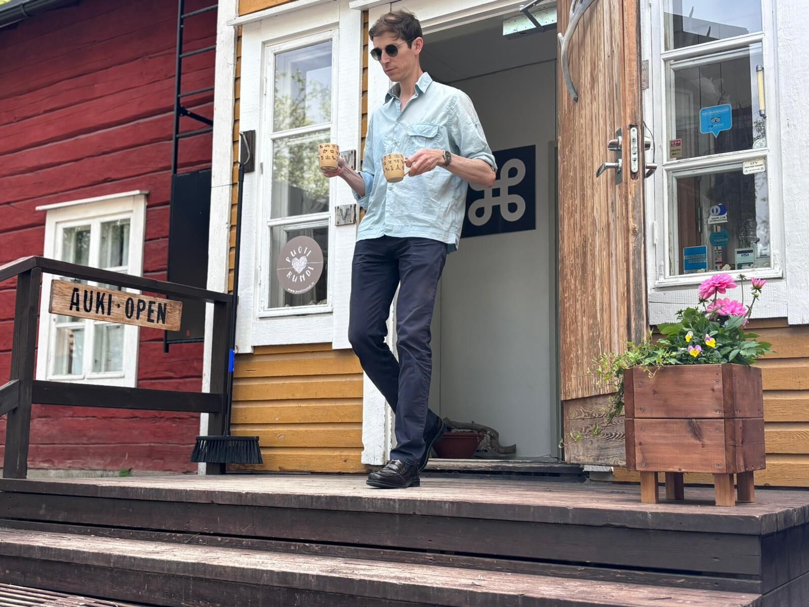 A person standing on a wooden porch with a sign reading 'AUKI OPEN' and a wooden planter with flowers.