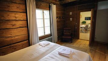 A wooden cabin room with two beds, a window with curtains, a chair, and a door leading to a dining area.