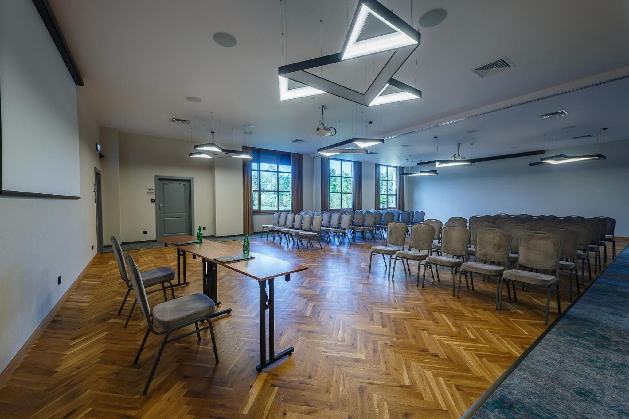 Un salón de conferencias con suelo de parqué labrado, filas de sillas, una mesa con una botella y ventanas amplias que dejan entrar luz natural.