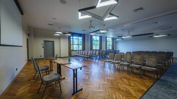 Un salón de conferencias con suelo de parqué labrado, filas de sillas, una mesa con una botella y ventanas amplias que dejan entrar luz natural.