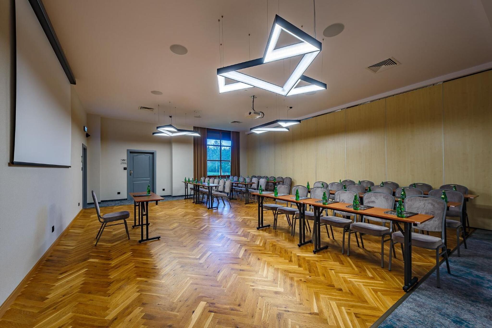 A conference room with a wooden herringbone floor, multiple tables with chairs, and modern geometric ceiling lights.