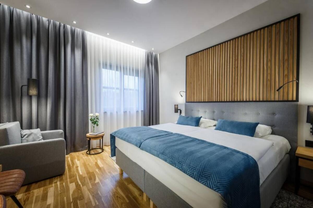 A modern hotel room with a large bed, a sofa, a small side table, and a window with curtains.