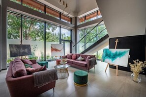 A modern living room with large windows, a pink sofa, a green ottoman, and an easel with a painting. - Hotel Prezydent (Inowlodz)