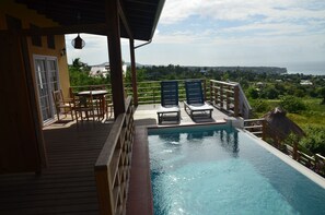 Área de piscina com deck de madeira, cadeiras de descanso e vista para o oceano e vegetação.
