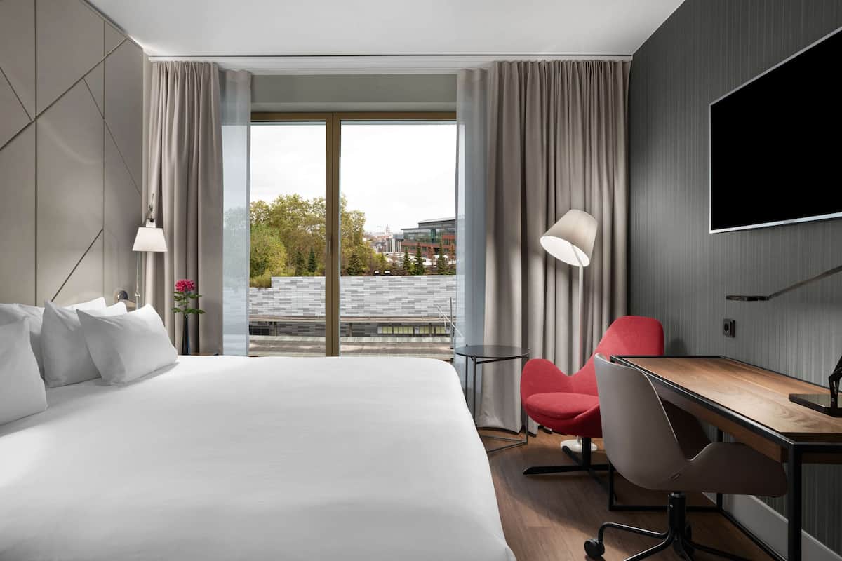 Une chambre d’hôtel dotée d’un grand lit, d’un bureau, d’une chaise, d’une télévision et offrant une vue sur la ville grâce à de grandes fenêtres.