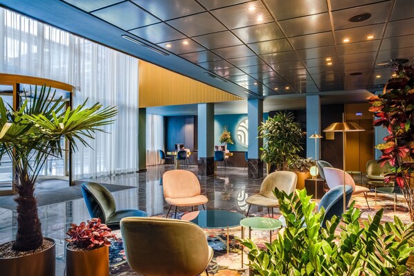 A modern lobby with a glass wall, seating areas, and potted plants. - NH Collection Antwerp Centre (Antwerp)