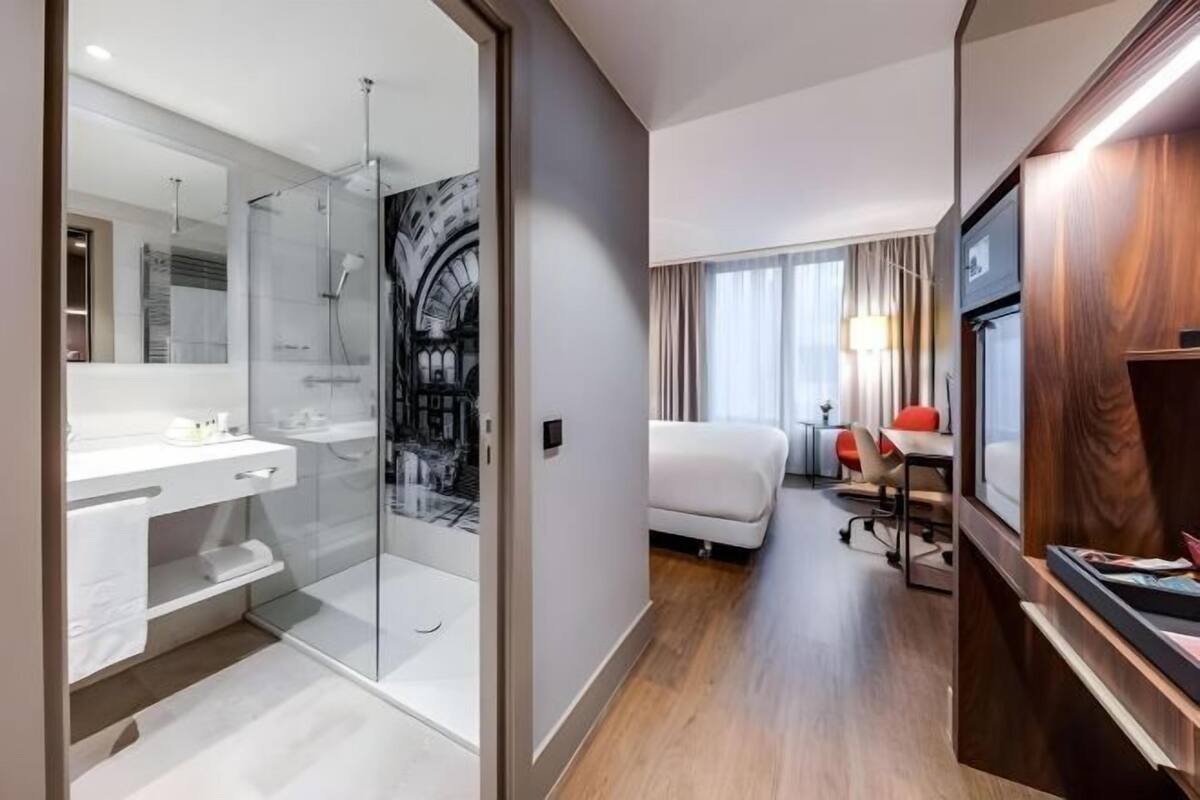 Une chambre d’hôtel moderne dotée d’une douche en verre, d’une coiffeuse blanche, d’un lit agrémenté d’une touche de rouge et d’un bureau en bois.