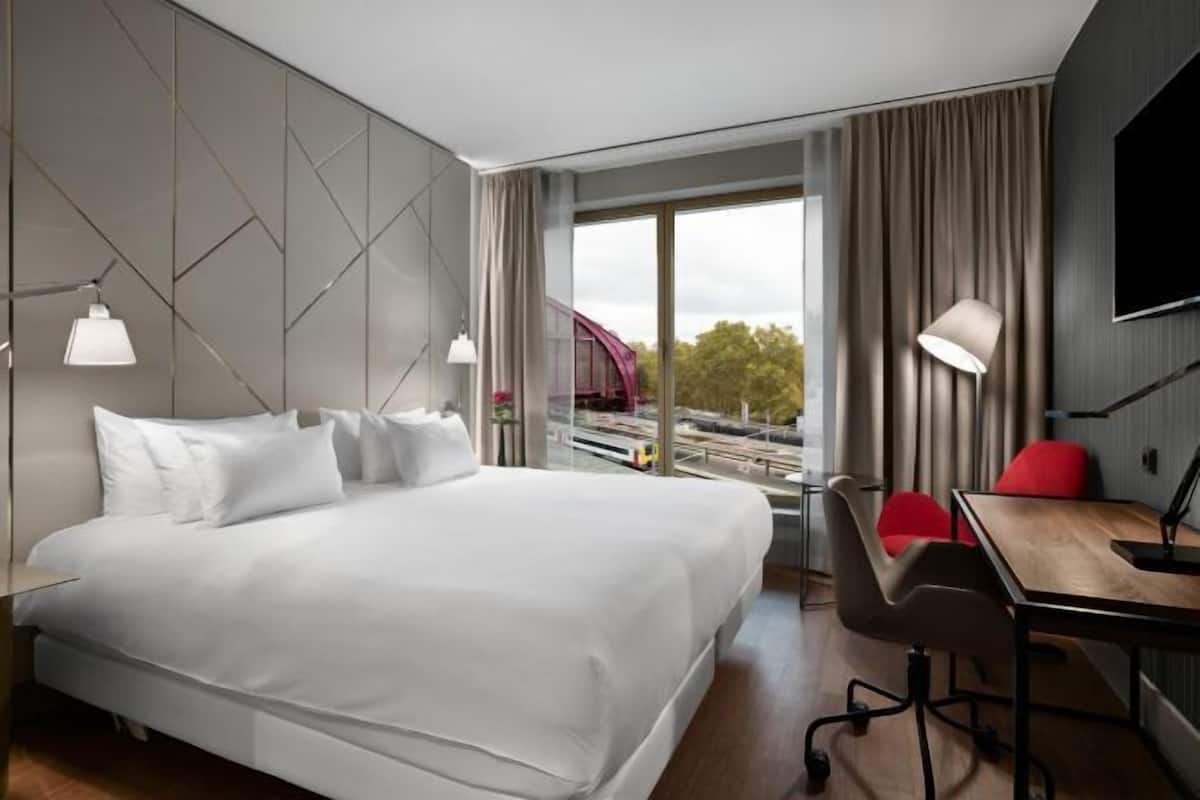 Une chambre d’hôtel moderne avec un grand lit, un bureau et une chaise. Une fenêtre offre une vue sur un pont et des arbres.