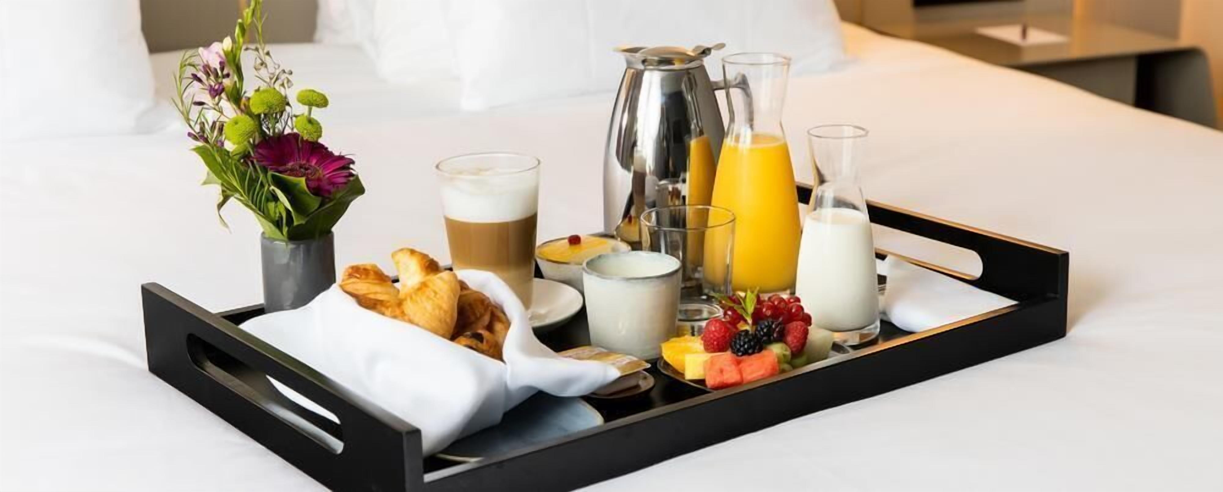A breakfast tray with croissants, coffee, orange juice, milk, and fresh fruits on a bed.