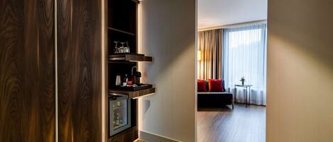 A hotel room with a wooden wardrobe, a small kitchenette, a seating area with a sofa and a table, and a window with curtains.