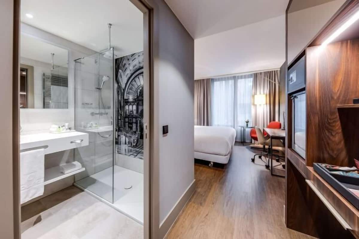 Une chambre d’hôtel moderne dotée d’une douche en verre, d’une coiffeuse blanche, d’un lit agrémenté d’une touche de rouge et d’un bureau en bois.