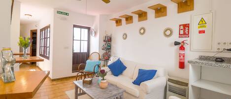 A well-lit living room with a white sofa, blue cushions, a wooden coffee table, and a ceiling fan. There is a bookshelf with books and decorative items, a fire extinguisher on the wall, and a sign indicating an exit.