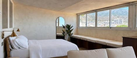 A bedroom with a bed, a bench, a round ottoman, and a view of a cityscape through large windows.