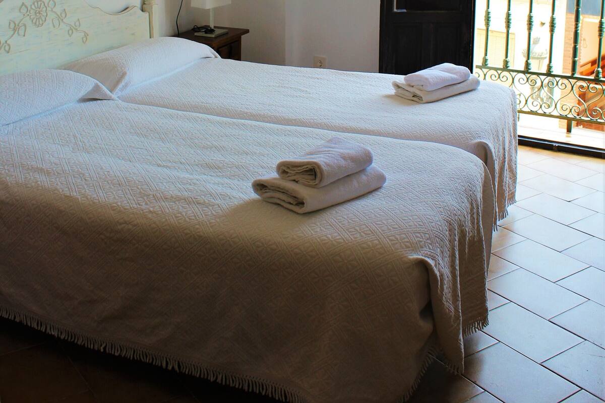 A hotel room with two beds, a nightstand with a lamp, and a balcony with a view.