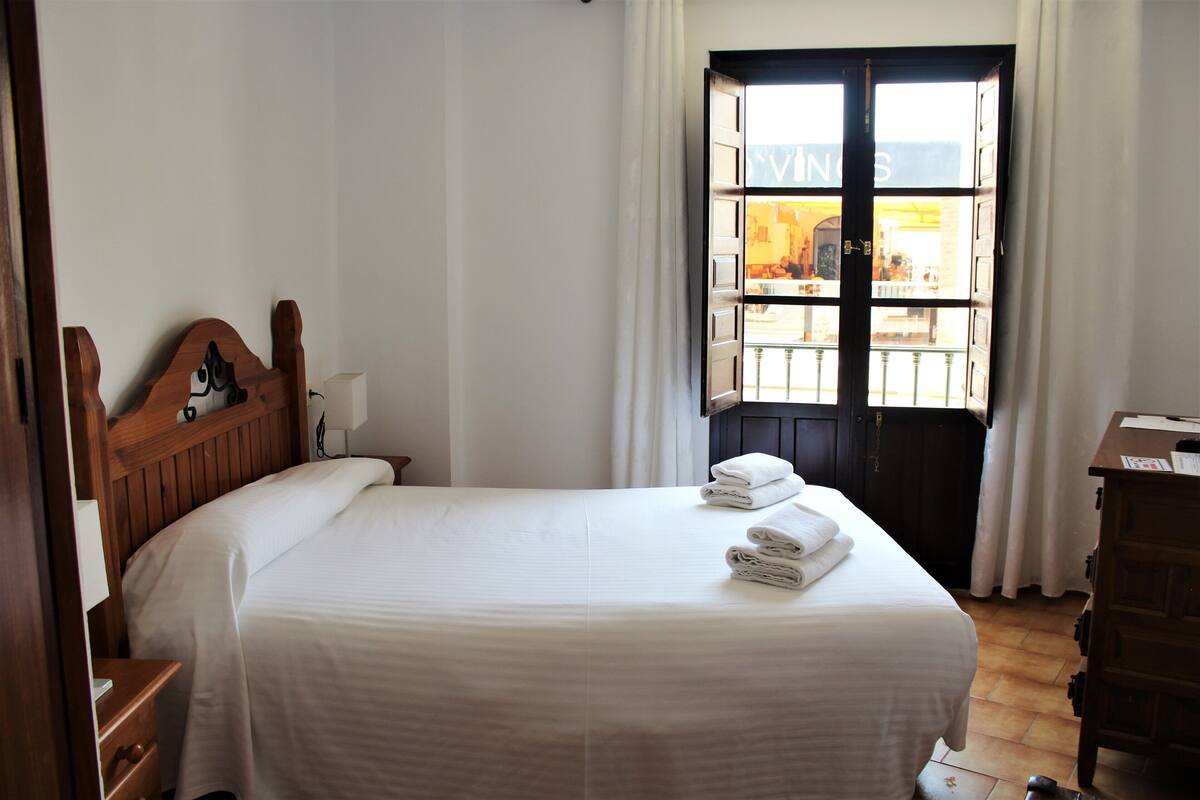 A neatly made bed with white linens and towels, a wooden headboard, a nightstand with a lamp, and a window with a view of a building outside.