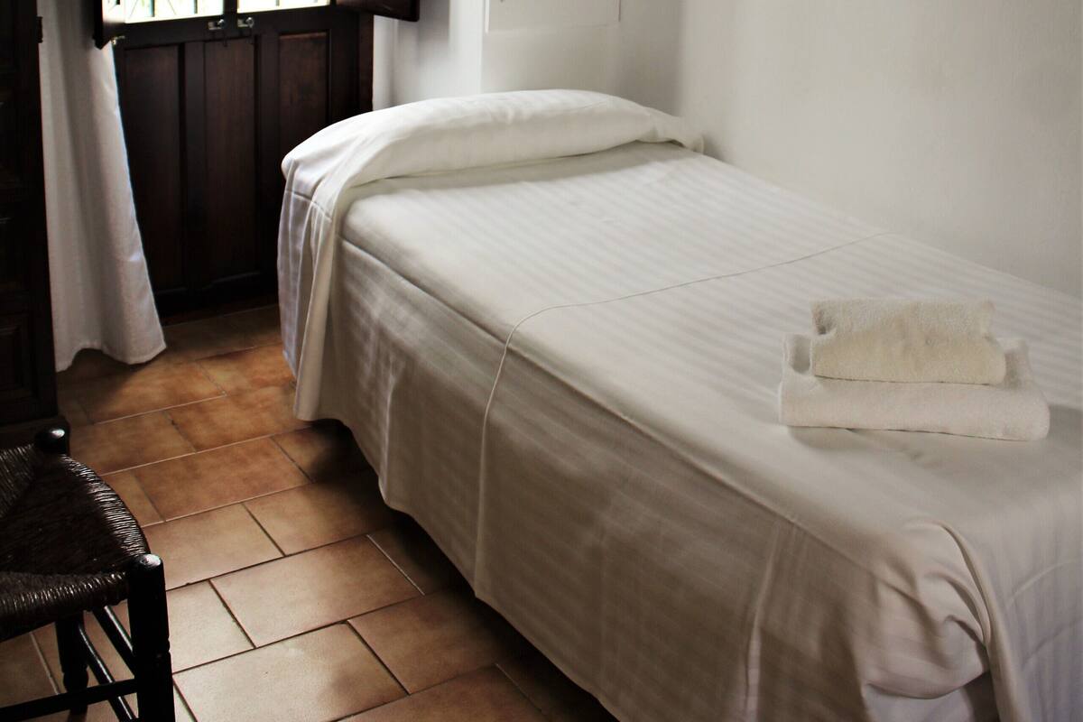 A single bed with a white bedspread and a pillow, a wooden chair, and a window with a view of greenery outside.