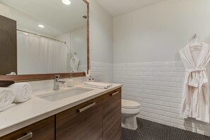 A modern bathroom with a white sink, vanity, and toilet. There is a robe hanging on the wall and towels on the counter.